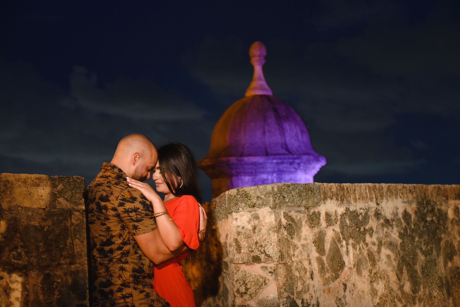 Couple embraces romantically at night near illuminated purple dome of historic fort wall under dramatic dark sky.