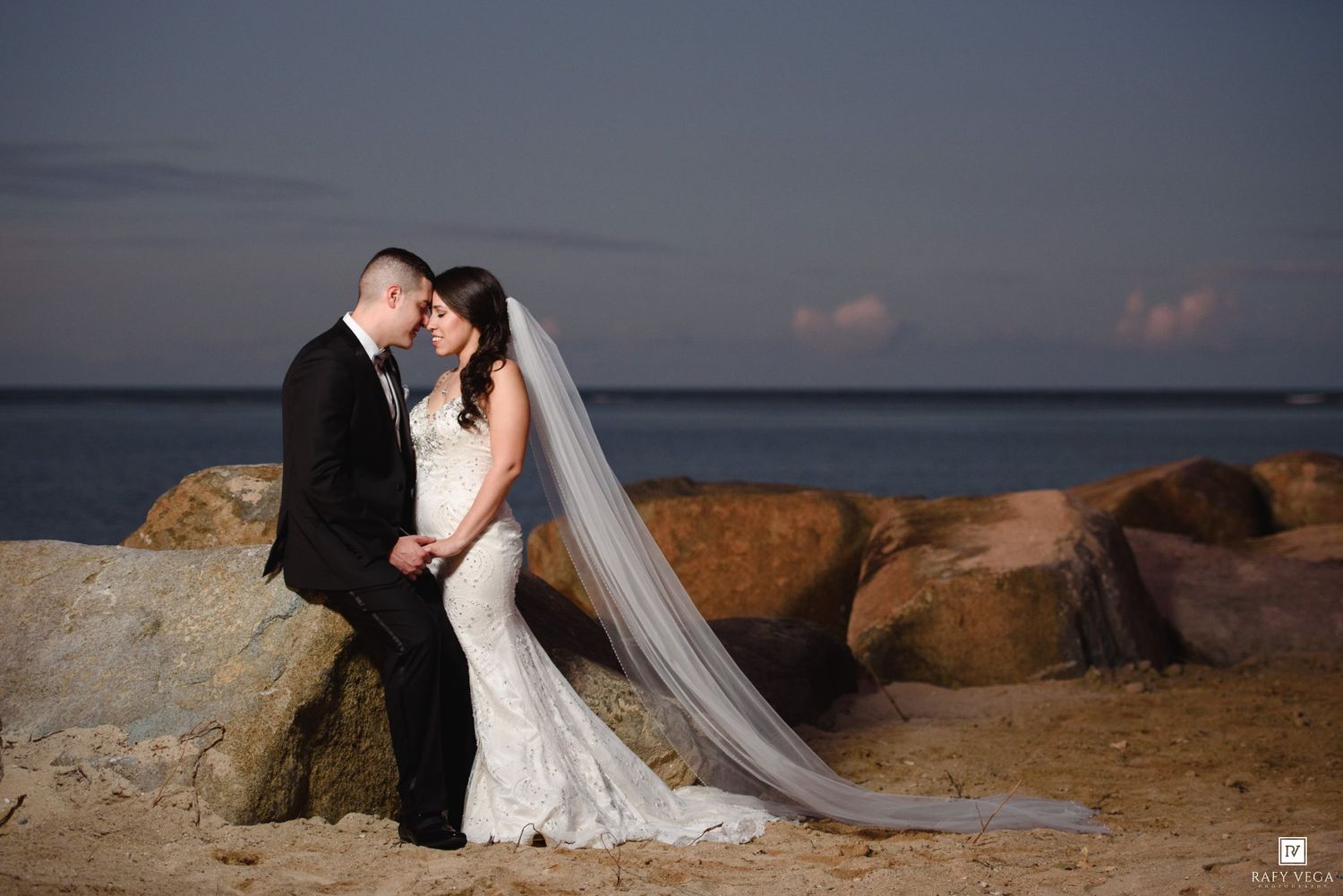 Bride and groom share a romantic moment by rocky seaside at sunset, her long veil flowing gracefully.