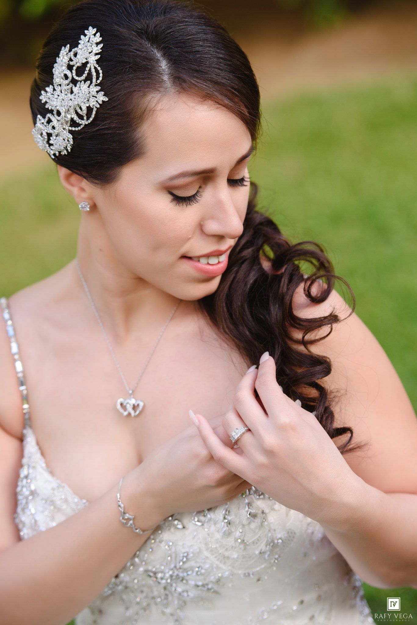 Elegant bride in beaded gown with crystal hair piece, double heart necklace, and diamond ring poses outdoors.