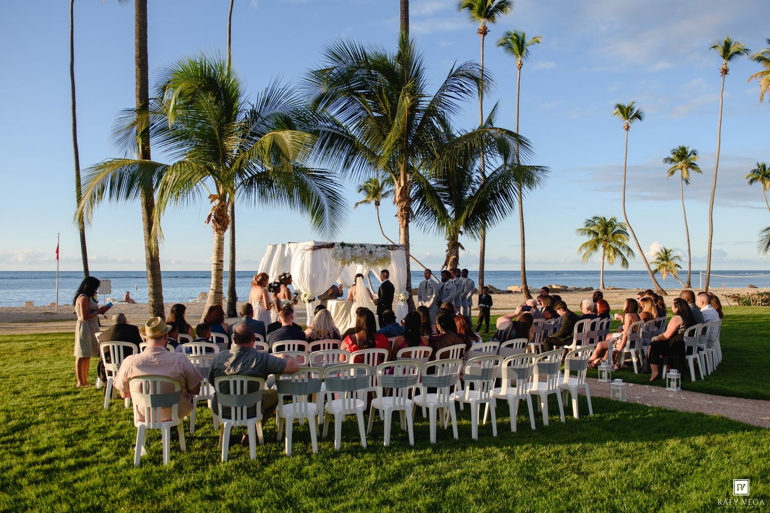 Outdoor beach wedding ceremony with guests seated on white chairs under tropical palm trees near the ocean.