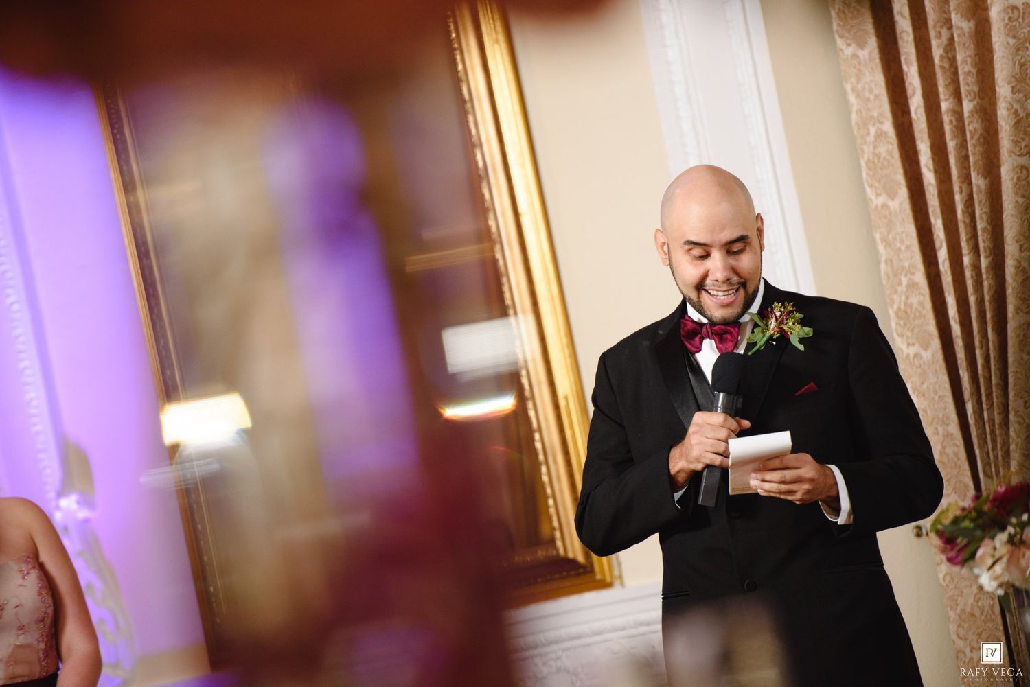 Bald groom in black tuxedo with red boutonniere reads wedding speech at elegant reception venue.