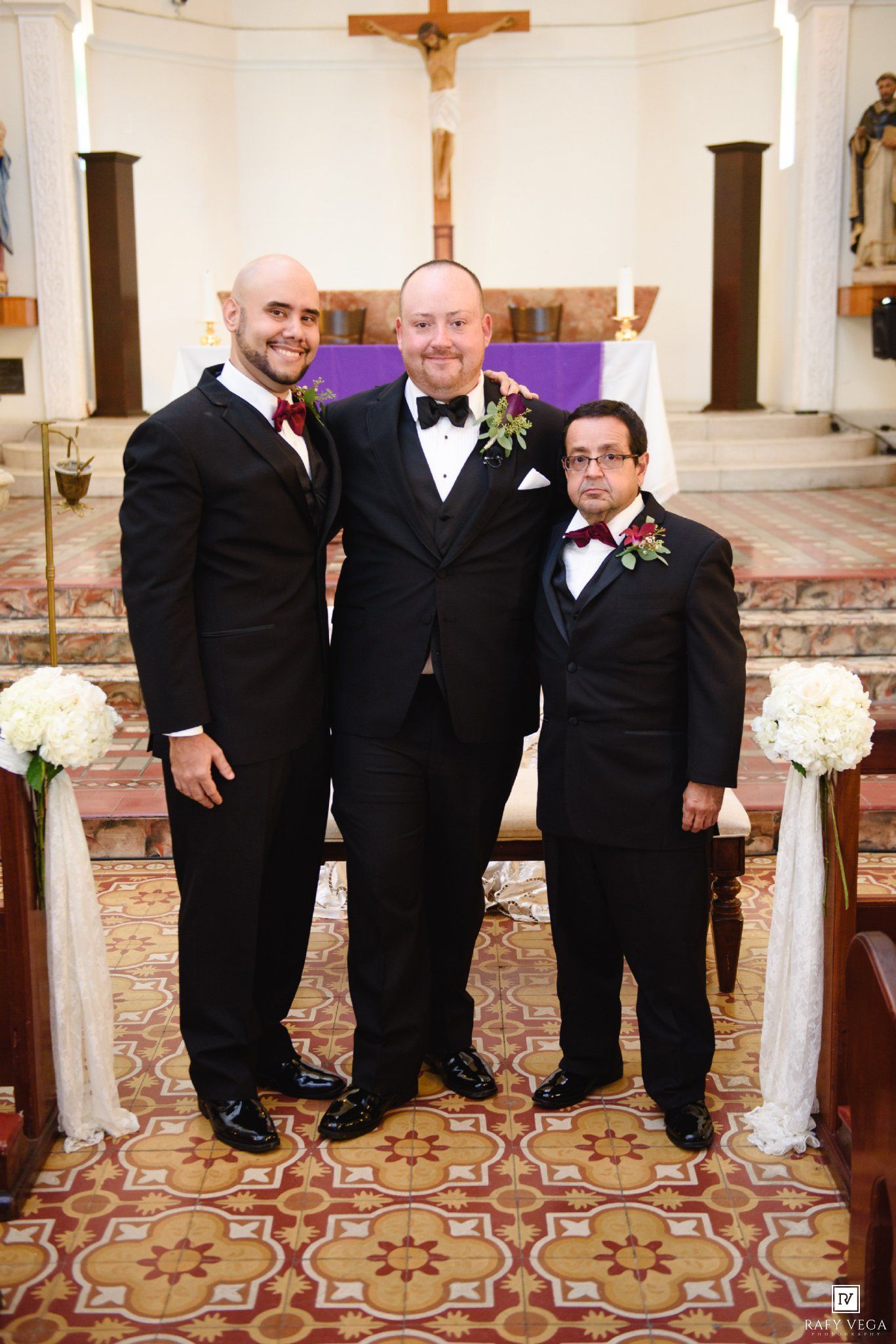 Three men in formal black tuxedos pose together inside a church decorated for a wedding ceremony.