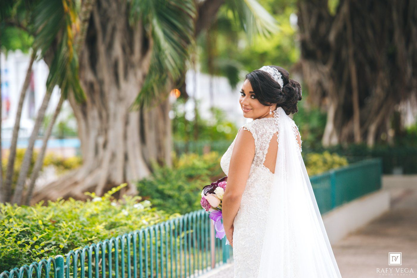 Bride in white lace gown and veil holding bouquet, posing outdoors near tropical trees and green fence.