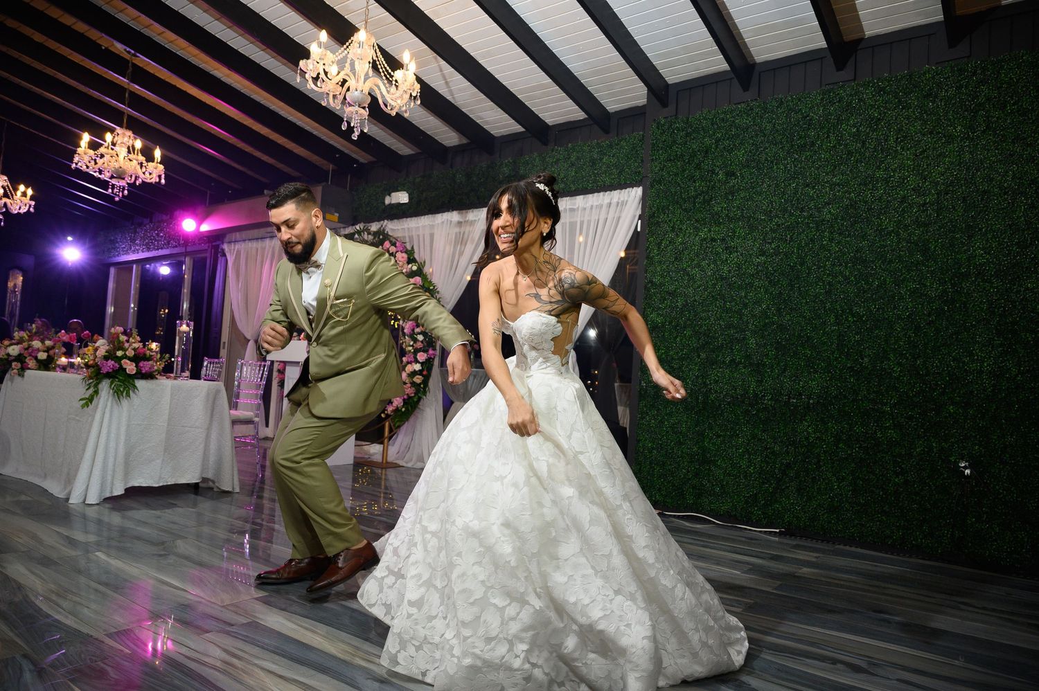 Bride in white ballgown and groom in olive suit dancing energetically at elegant wedding reception with green floral wall backdrop.