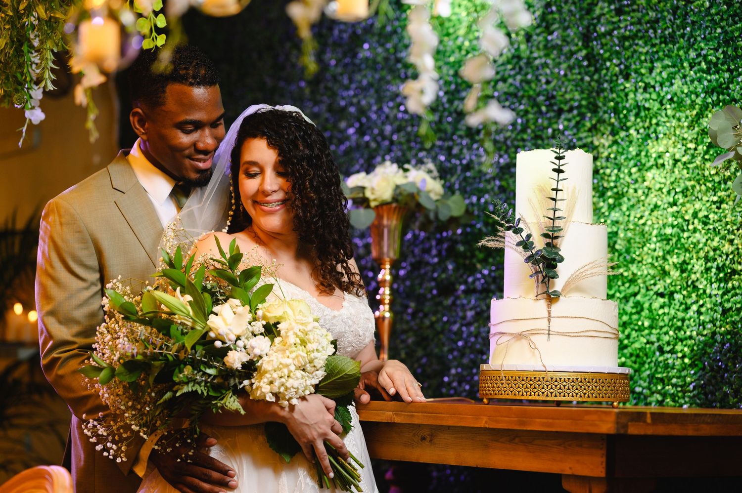 Interracial couple cutting wedding cake, bride holding white bouquet, green floral wall backdrop.