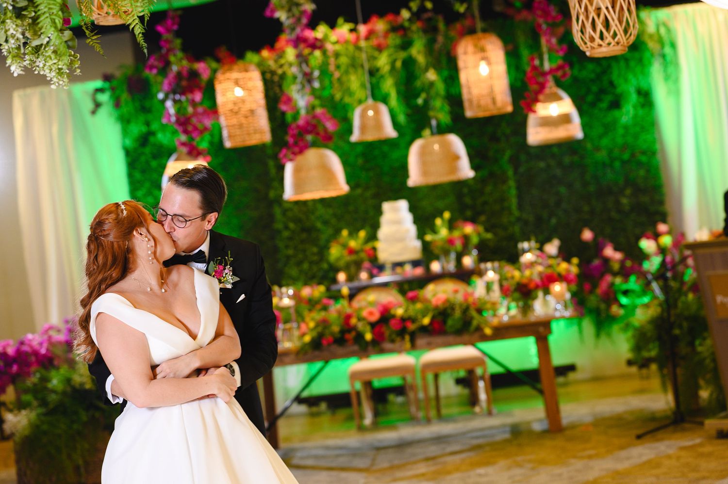 Bride and groom share a romantic kiss at elegant wedding reception with floral backdrop and hanging lanterns.