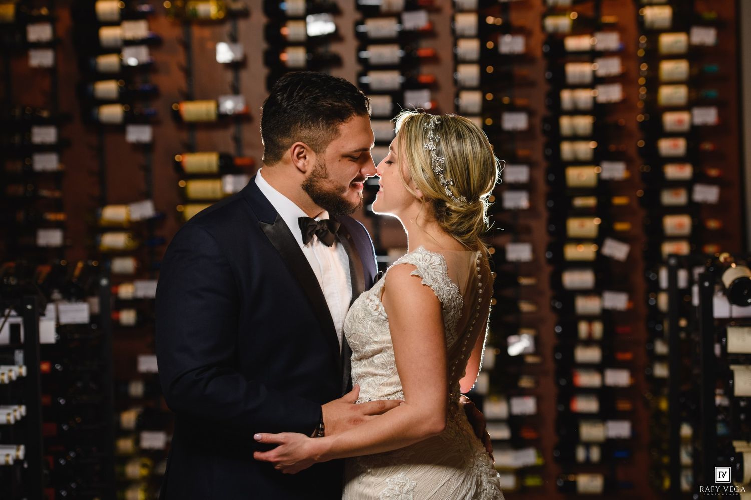 Bride and groom share a romantic moment in a wine cellar, surrounded by walls of wine bottles.