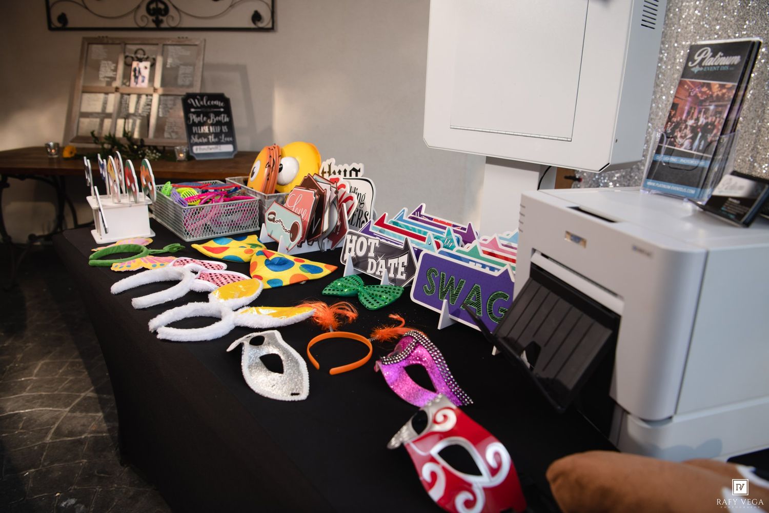 Photo booth props and masks displayed on a black table with a printer at a party or wedding event.