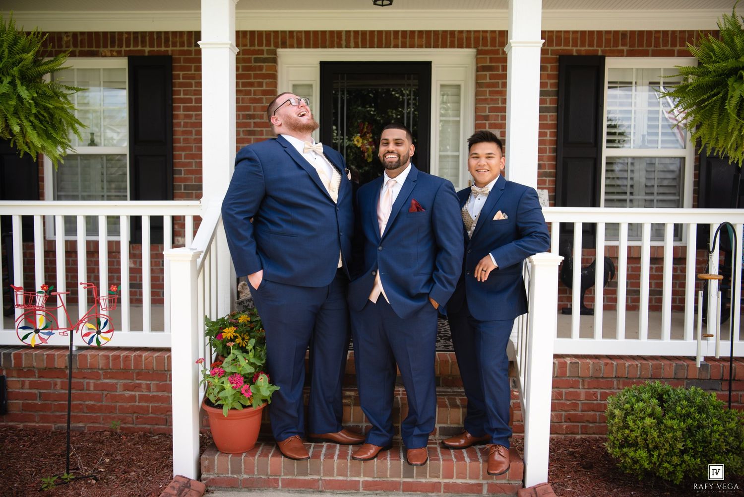 Three groomsmen in navy blue suits posing on brick porch steps of a brick house for a wedding photo.
