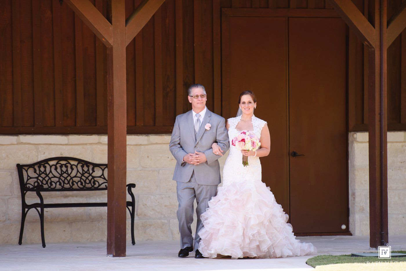 Bride in ruffled white gown walks with escort under wooden pavilion at outdoor wedding ceremony.