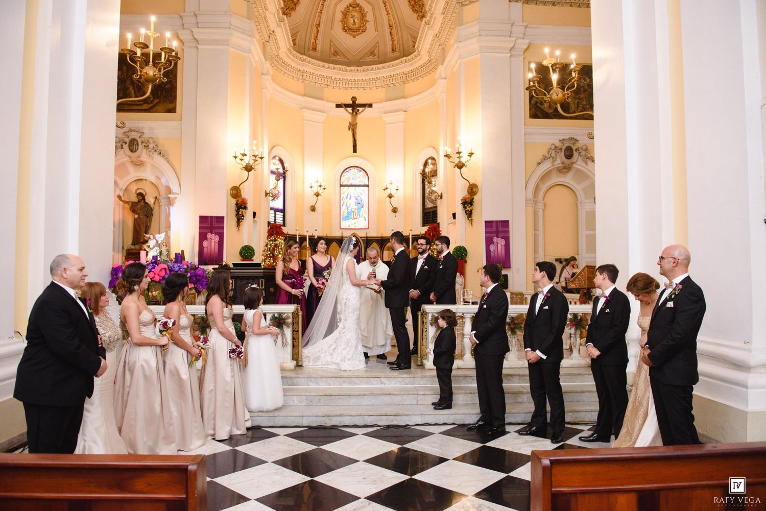Wedding party stands at church altar with checkered marble floors, bridesmaids in blush gowns and groomsmen in black suits.
