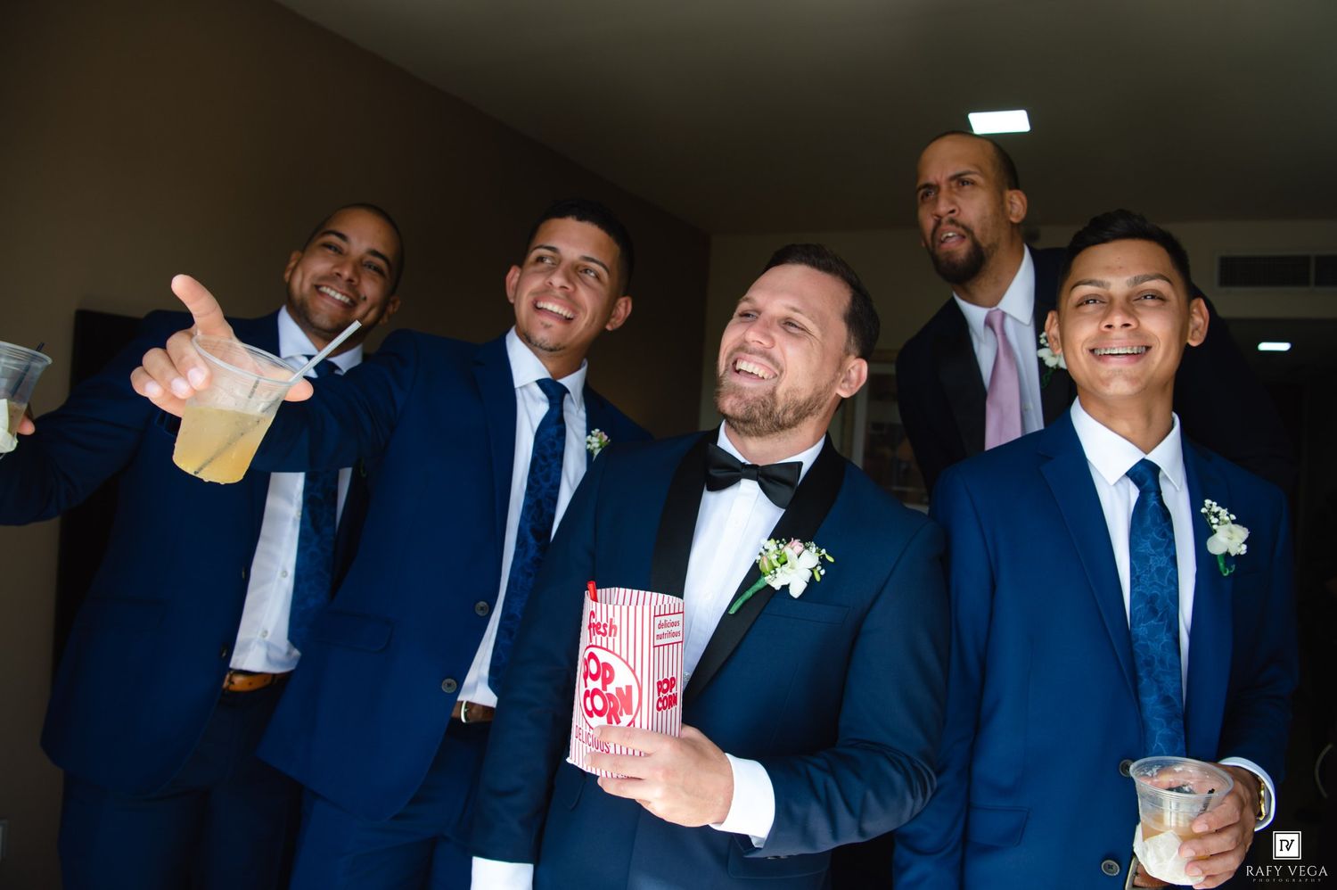 Groom in black tuxedo with groomsmen in navy suits celebrating together before wedding ceremony.