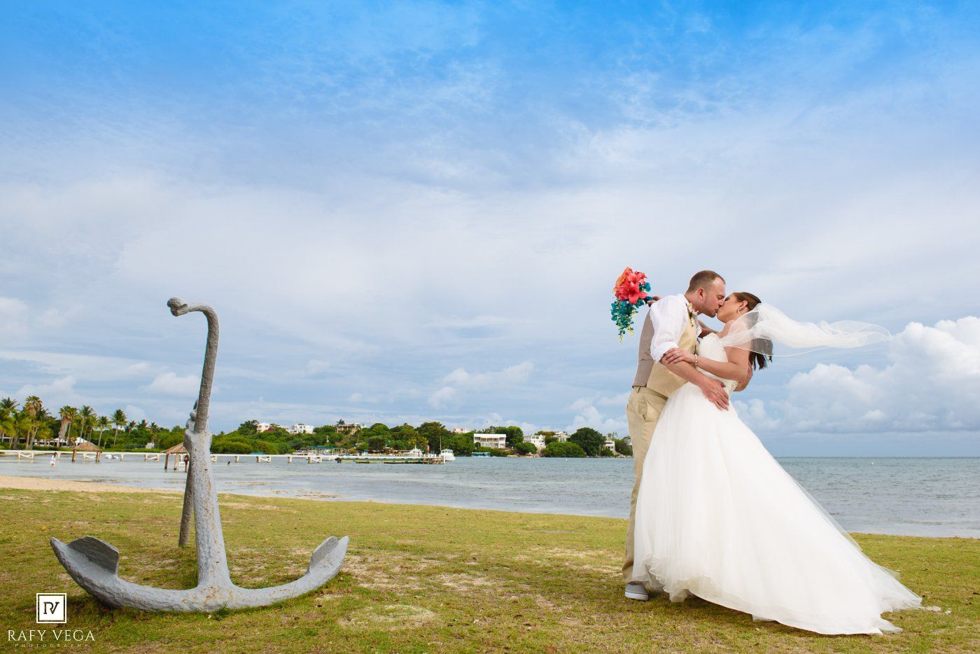 Bride and groom kissing near a large anchor on a tropical beach with blue skies and ocean views.