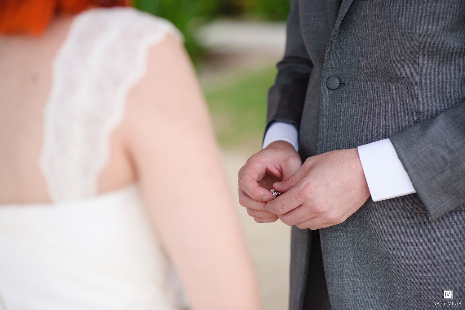 Groom in gray suit holds bride's hand during outdoor wedding ceremony.