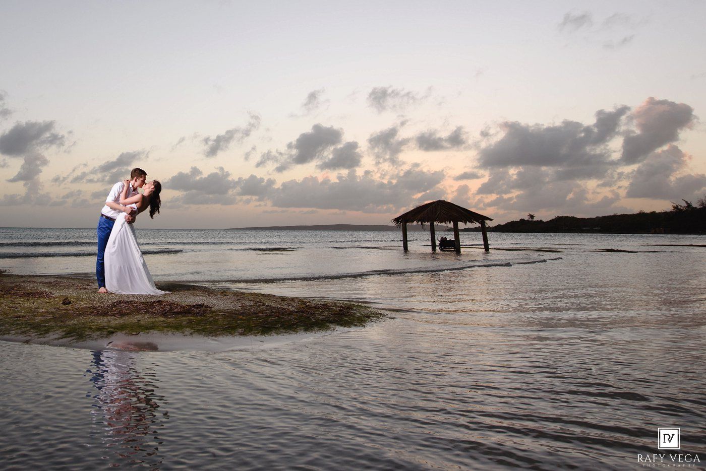 Couple sharing a romantic kiss on a tropical beach at sunset with a thatched gazebo in the background.