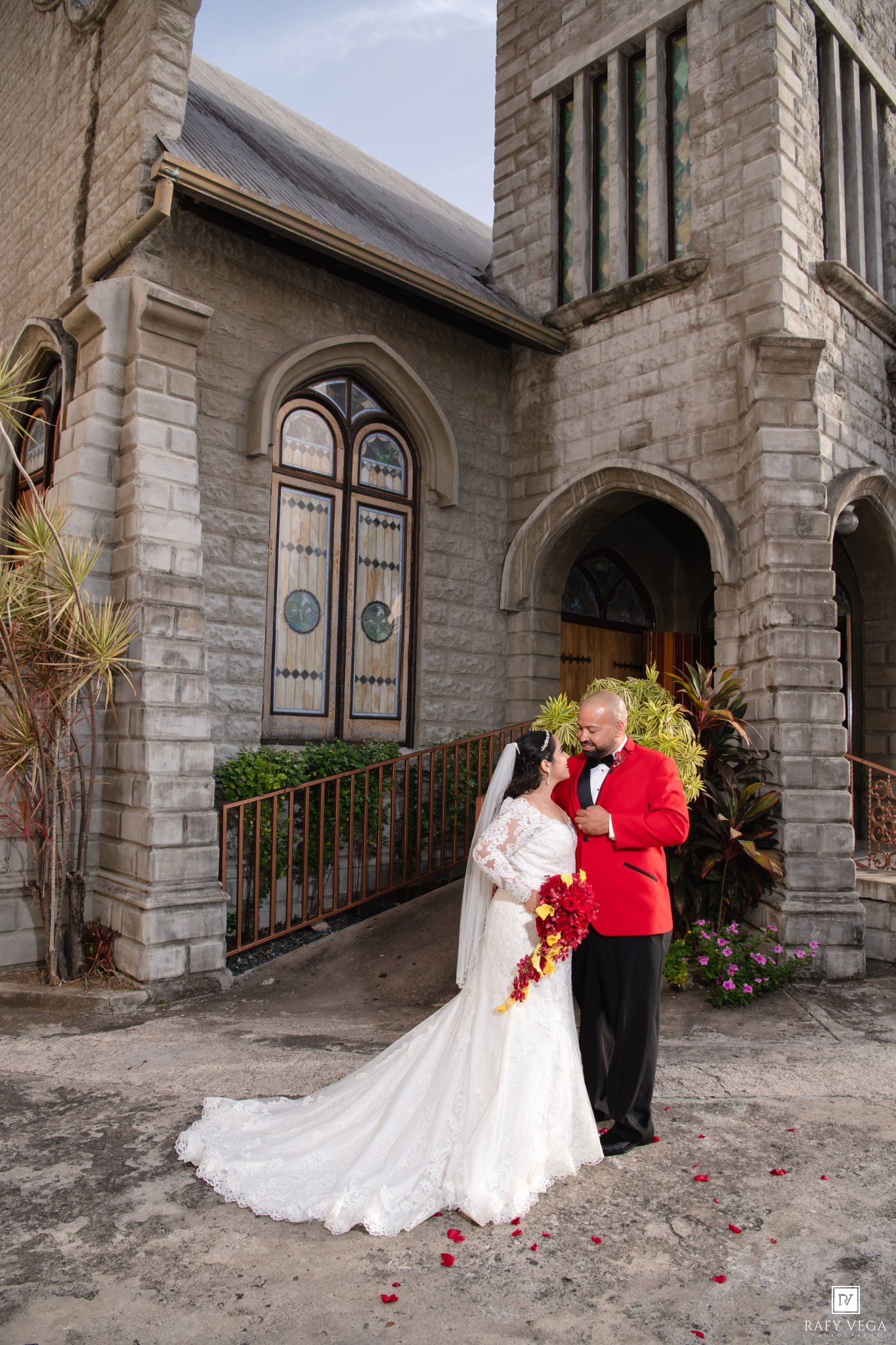 Bride in white gown and groom in red tuxedo share a romantic moment outside a stone church on their wedding day.