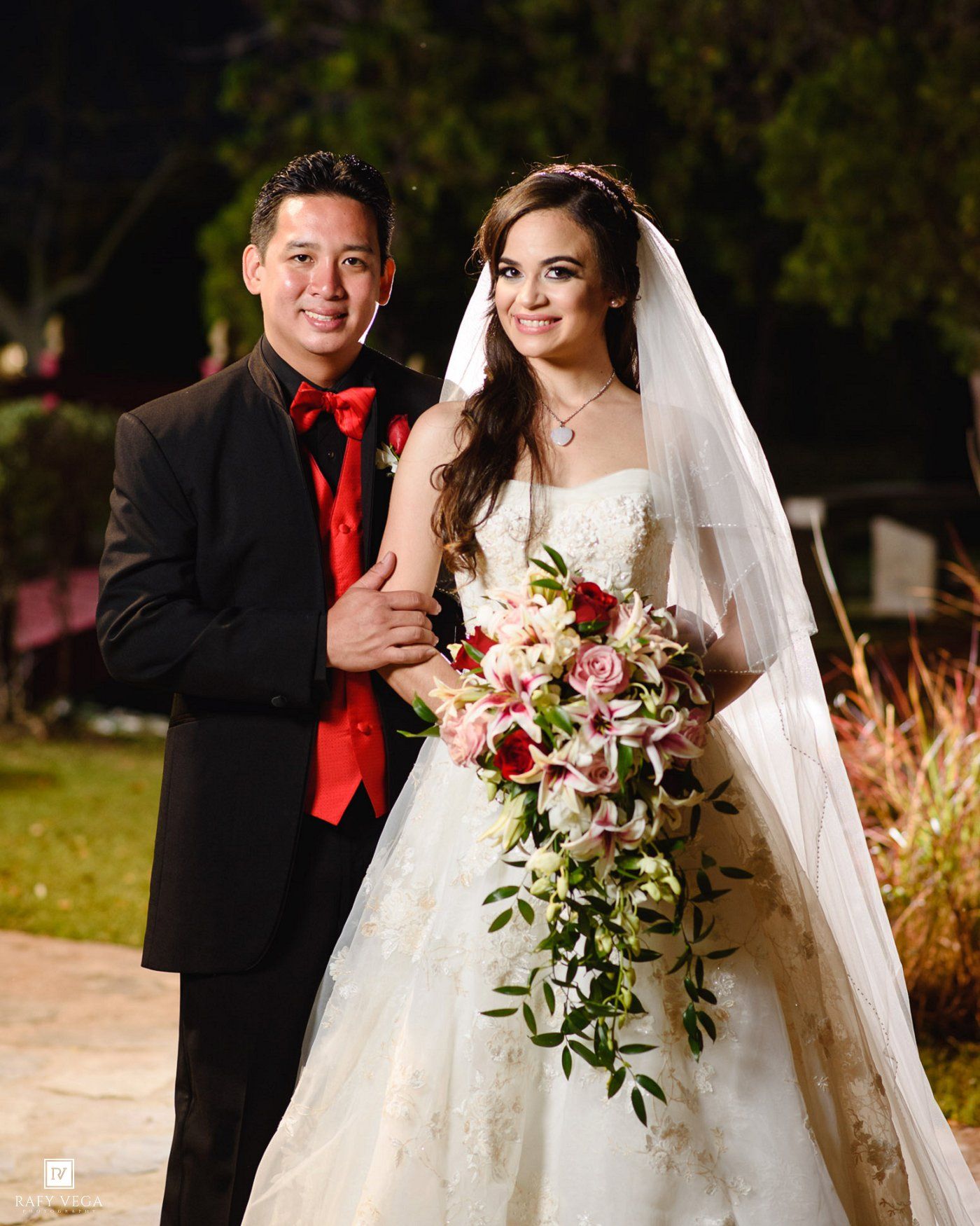 Bride and groom posing outdoors at night, groom in black tuxedo with red tie, bride in white gown holding floral bouquet.