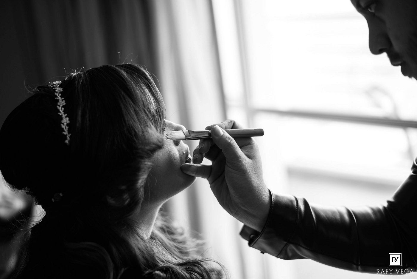 Black and white photo of a bride having makeup applied by a makeup artist before her wedding ceremony.