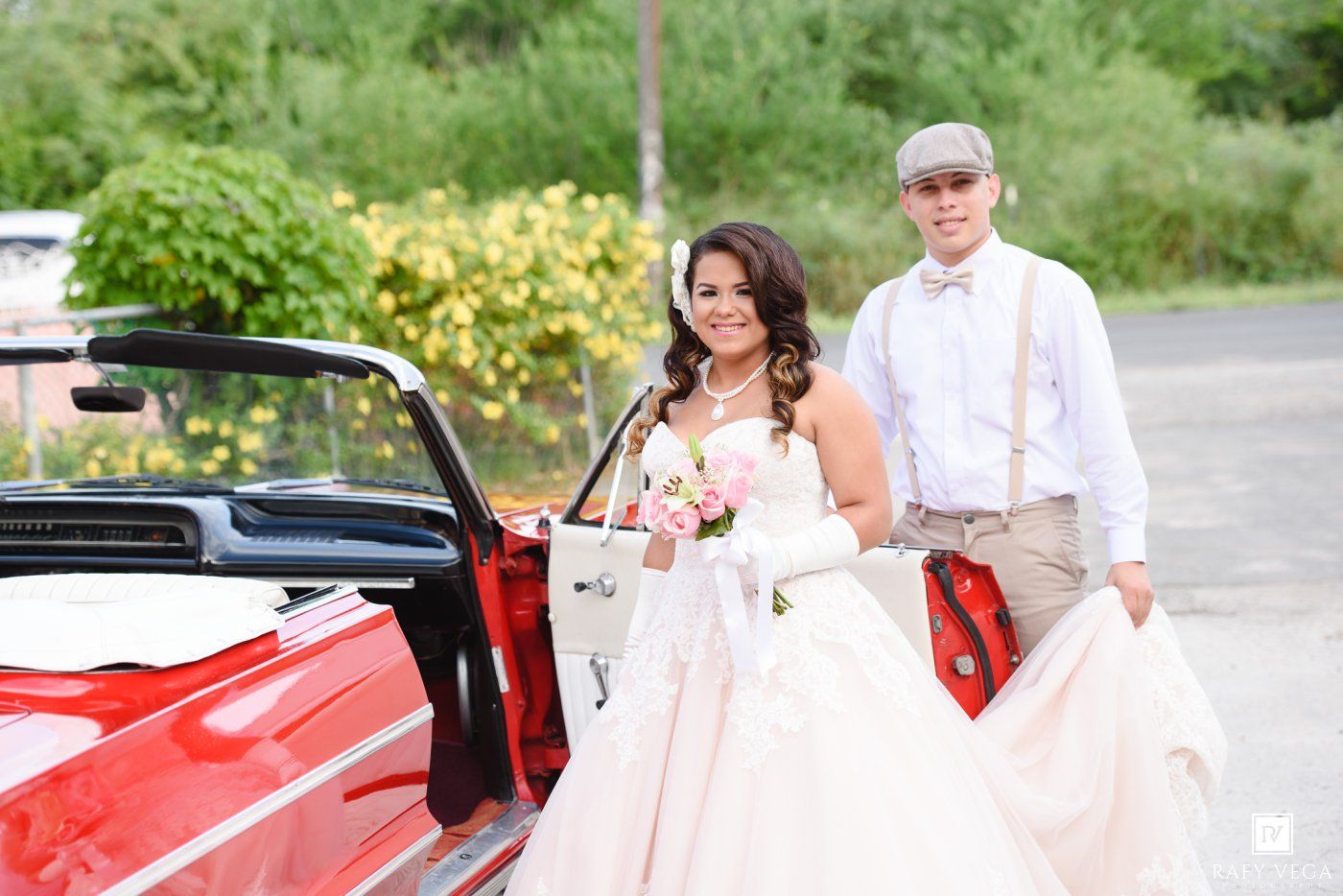 Bride in white gown and groom in suspenders pose beside a classic red convertible car outdoors.