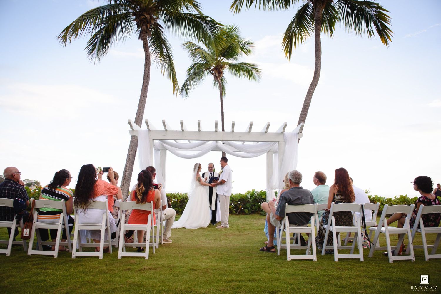 Outdoor beach wedding ceremony with white chairs, floral arch, palm trees, and guests gathered on a tropical lawn.