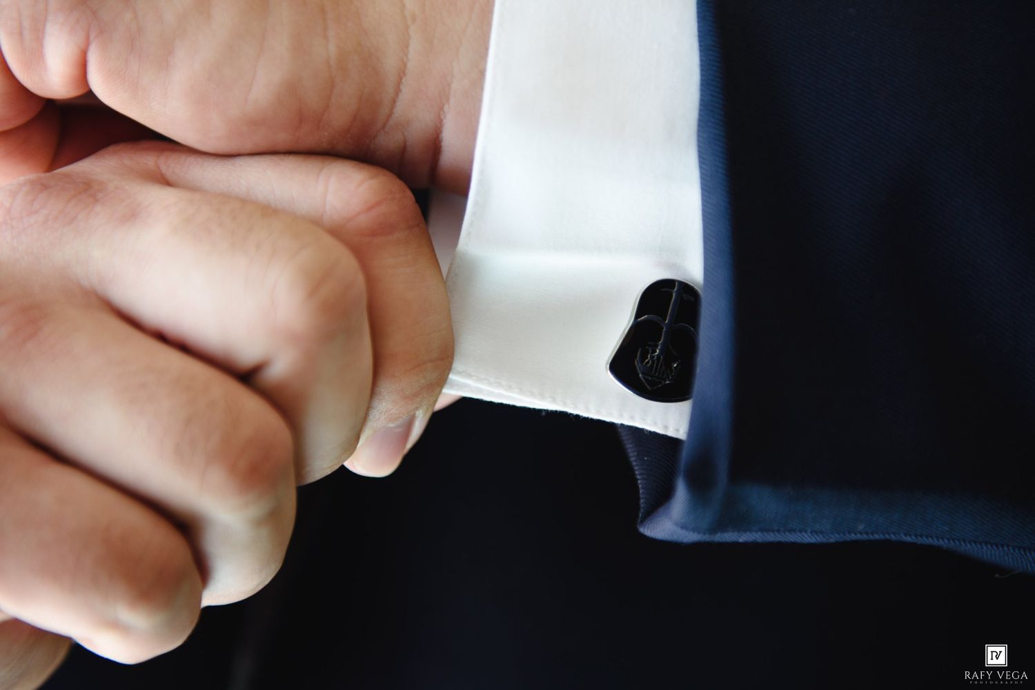 Close-up of a man fastening a black cufflink on a white dress shirt sleeve with a navy suit jacket.