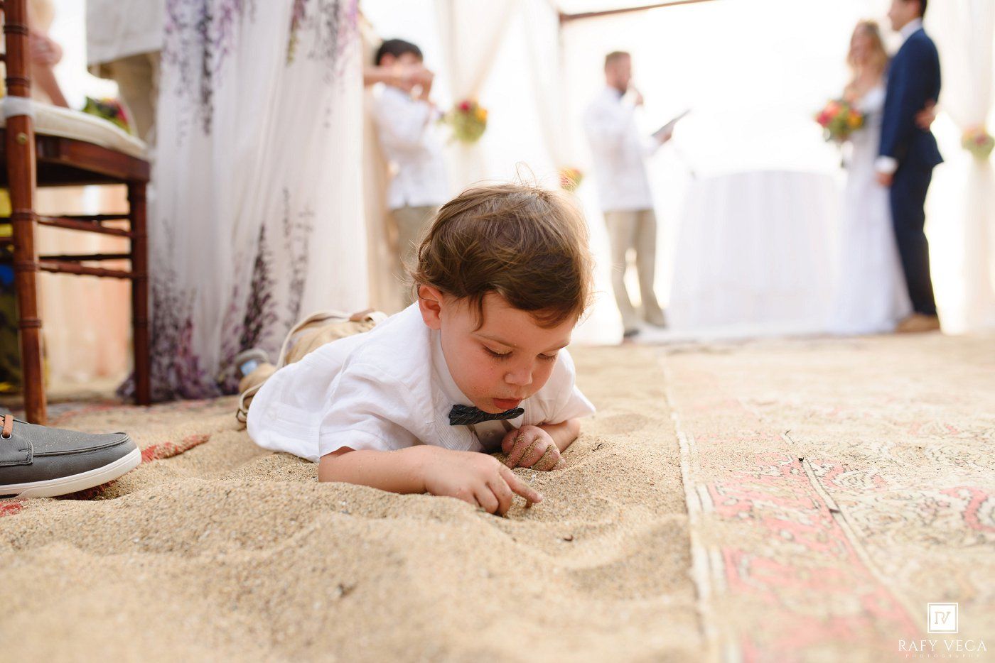 Young ring bearer boy playing in sand during outdoor beach wedding ceremony with bride and groom in background.