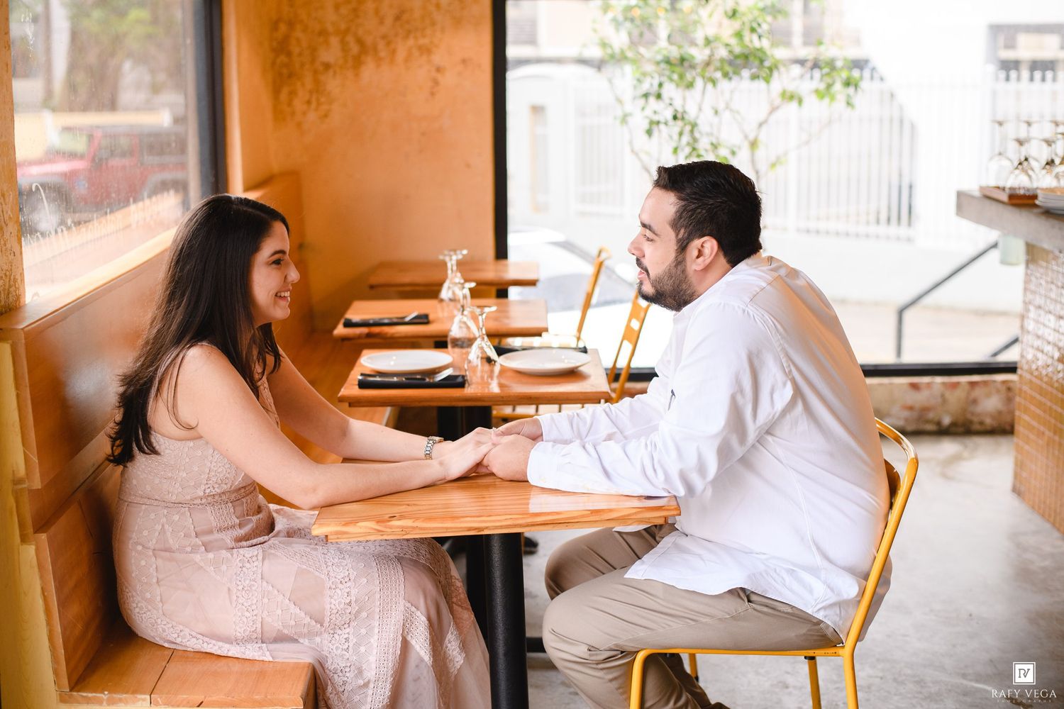 Couple holding hands and talking at a wooden restaurant table by a window in a warm, cozy dining setting.