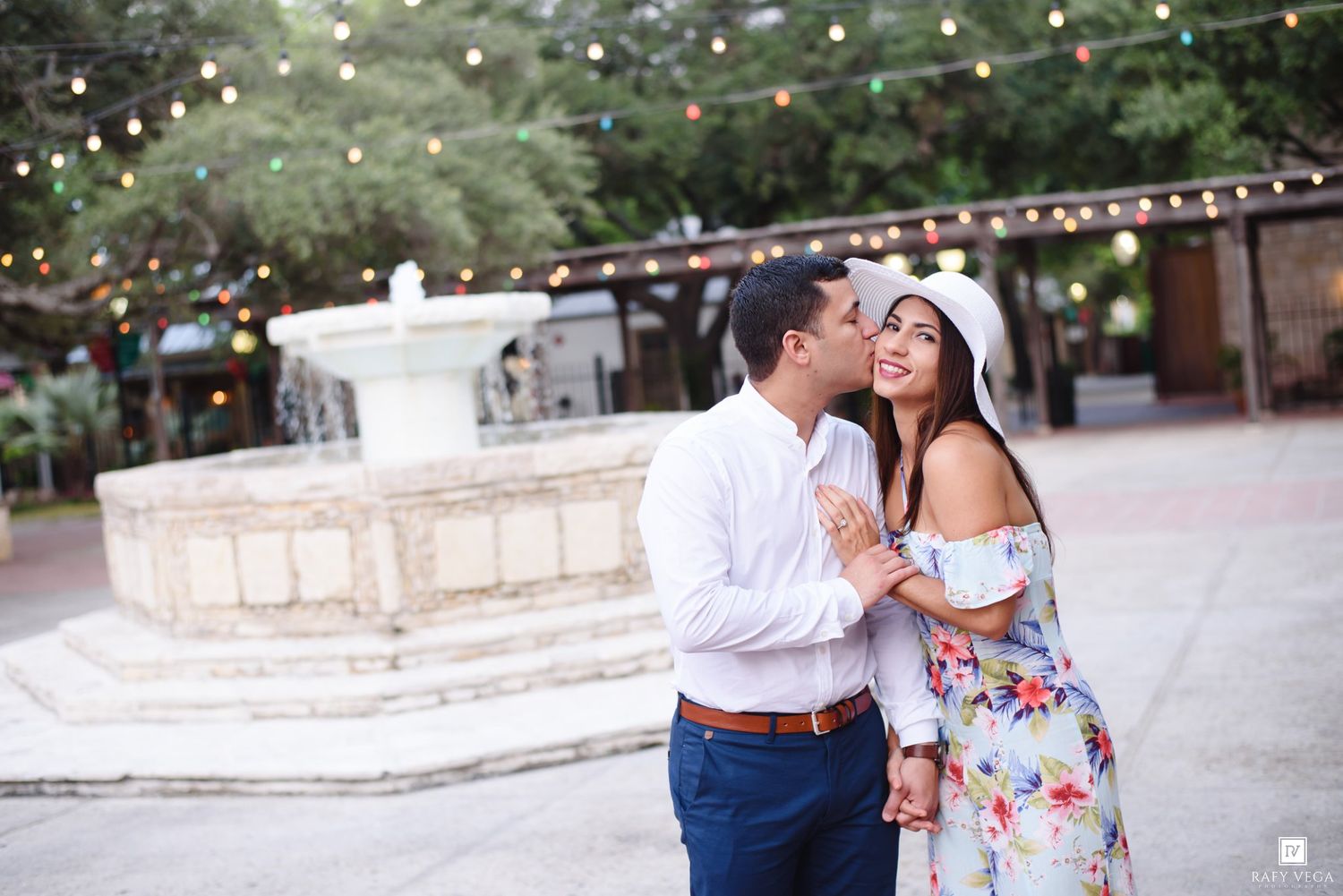 Couple sharing a romantic kiss near a white fountain with string lights at an outdoor venue.