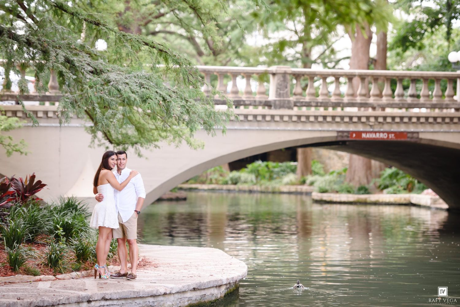 Couple embracing in white outfits by a scenic river walk bridge surrounded by lush green trees.
