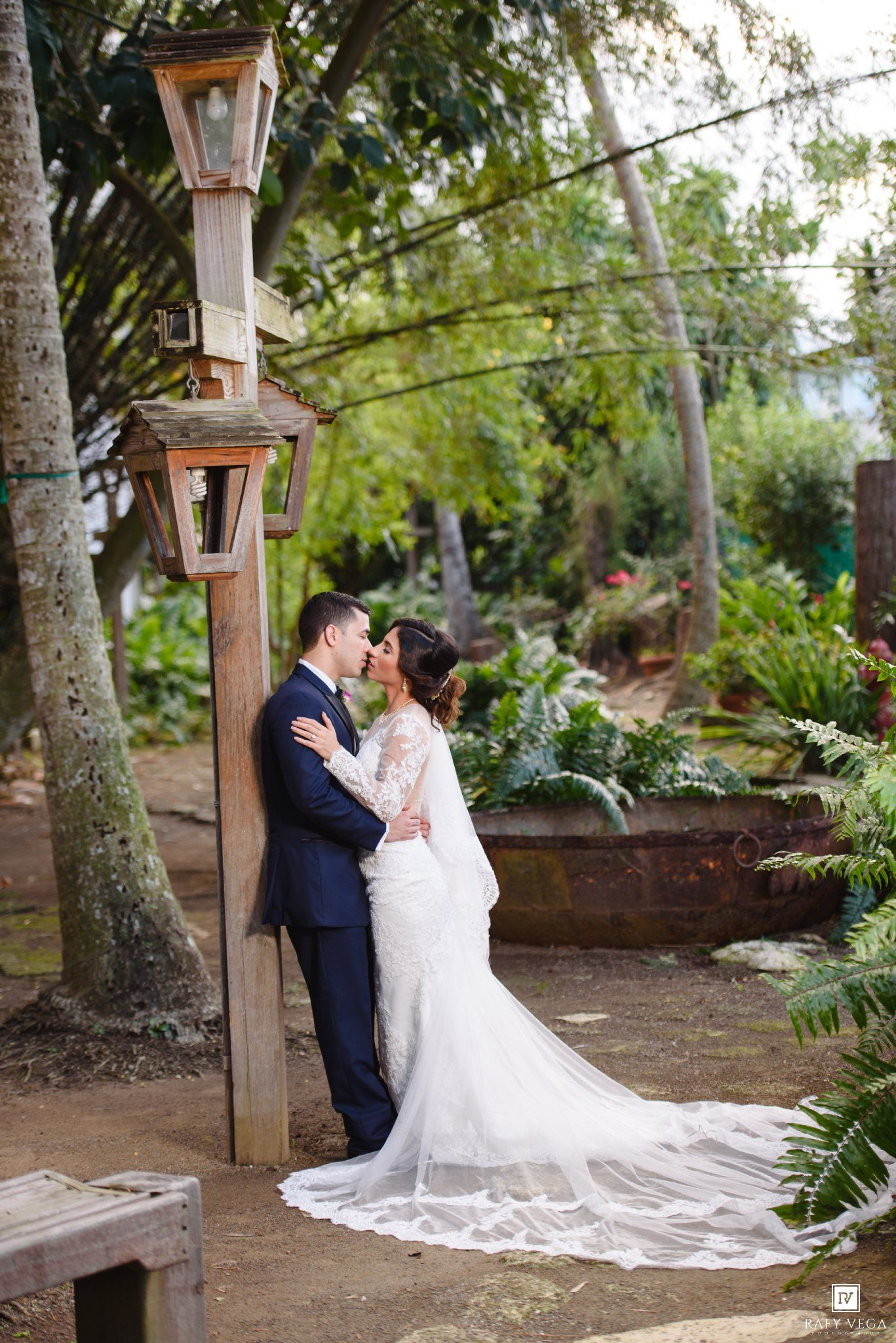 Bride and groom share a romantic kiss in a lush garden setting, bride wearing a long lace wedding gown with train.