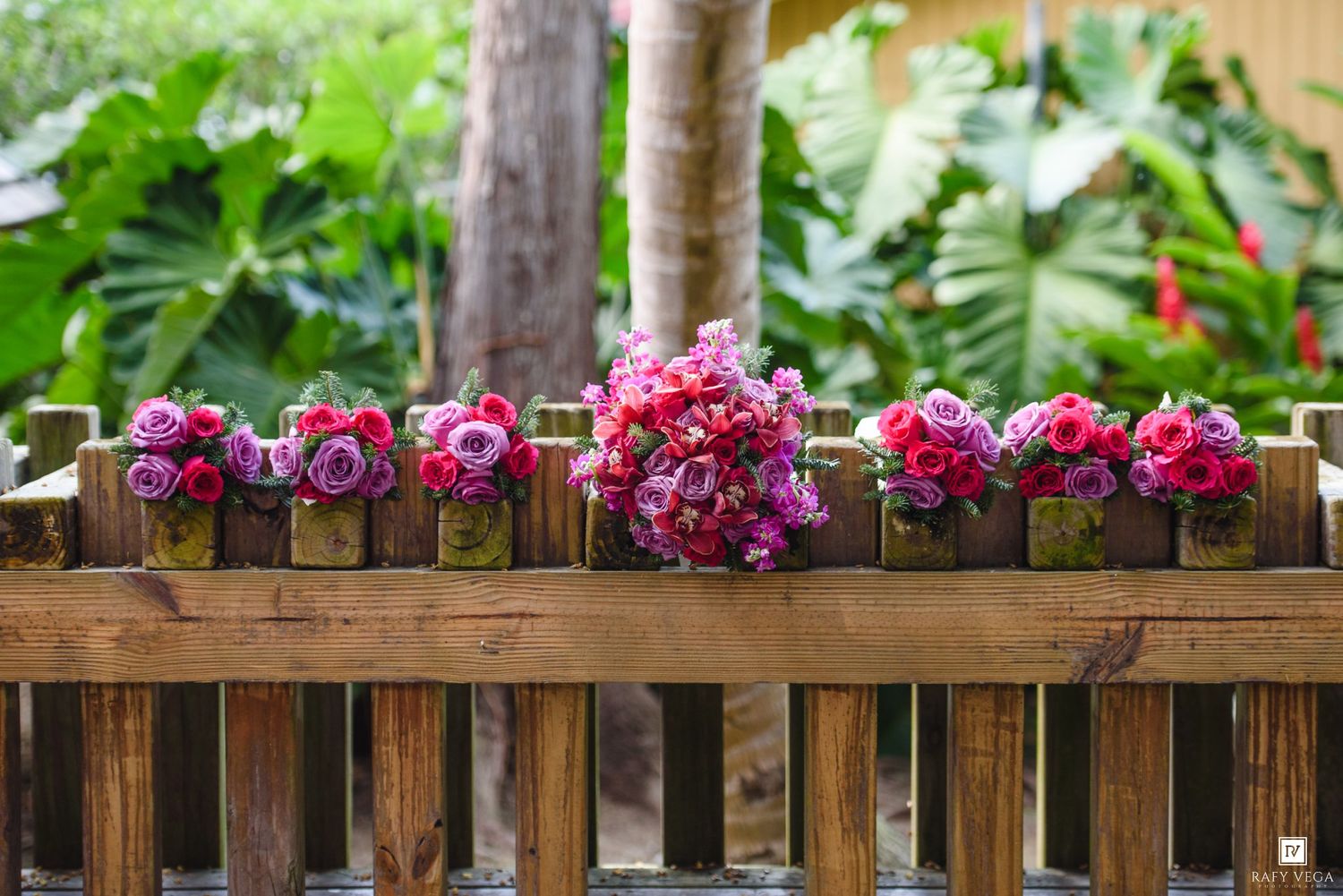 Red and pink floral bouquets displayed on a rustic wooden fence surrounded by lush tropical greenery.