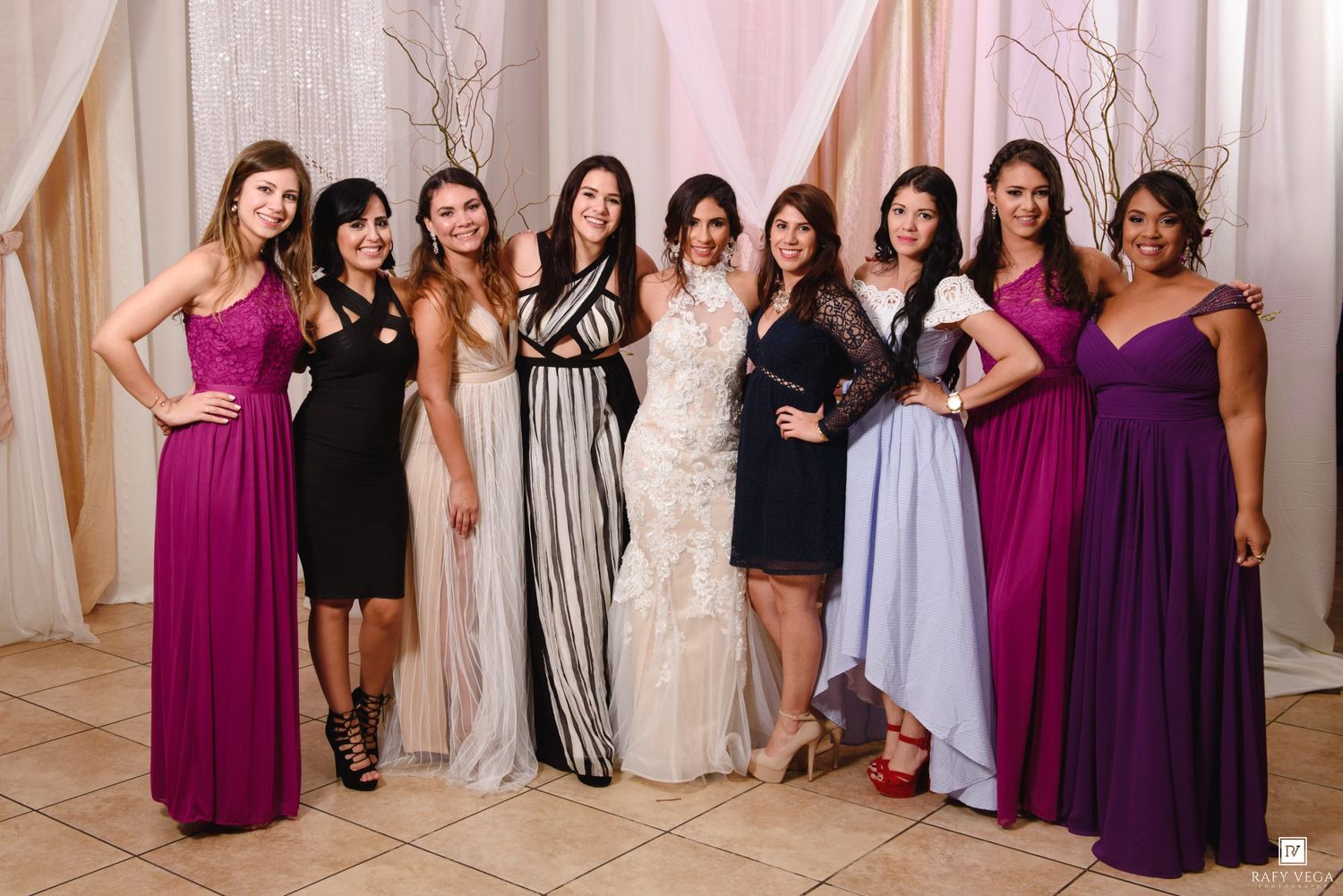 A group of nine women in elegant formal gowns pose together at a wedding celebration in a decorated venue.