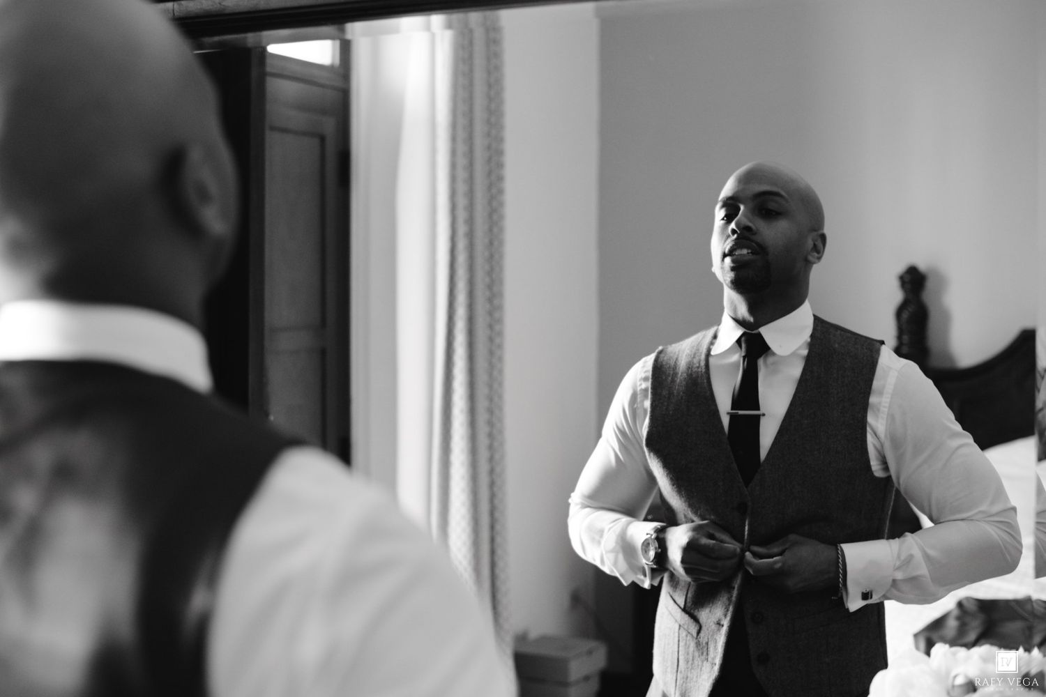 Black and white photo of a well-dressed man in a vest and tie adjusting his outfit in front of a mirror.
