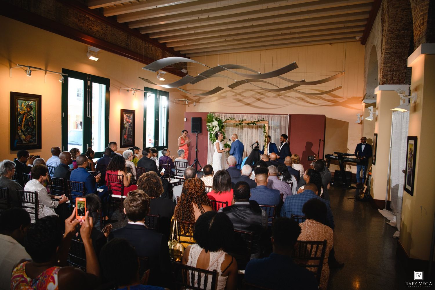 Indoor wedding ceremony in an art gallery with guests seated, couple at altar, decorative walls, and elegant chandelier lighting.