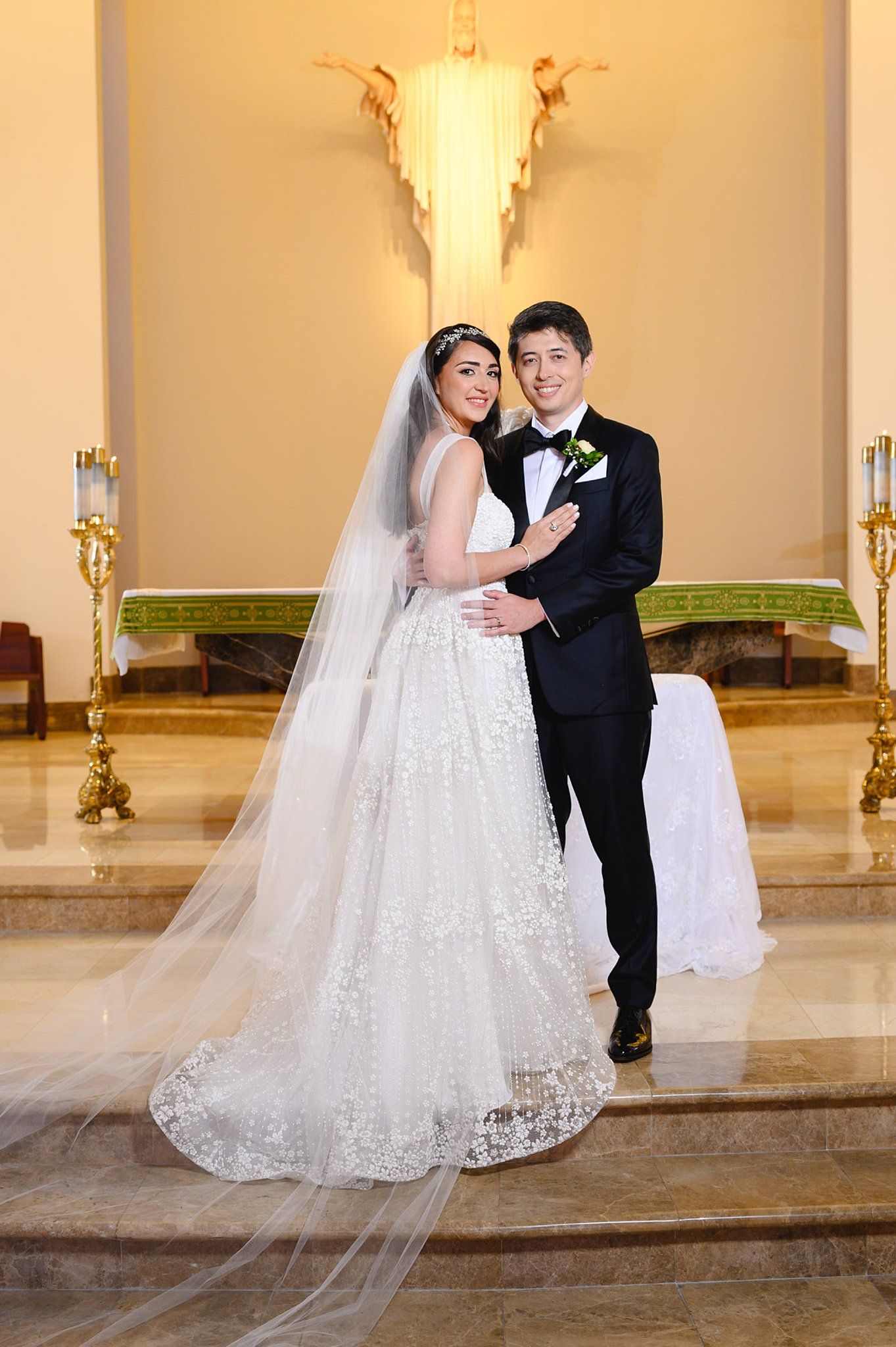 Bride in lace gown and veil poses with groom in black tuxedo at church altar on their wedding day.