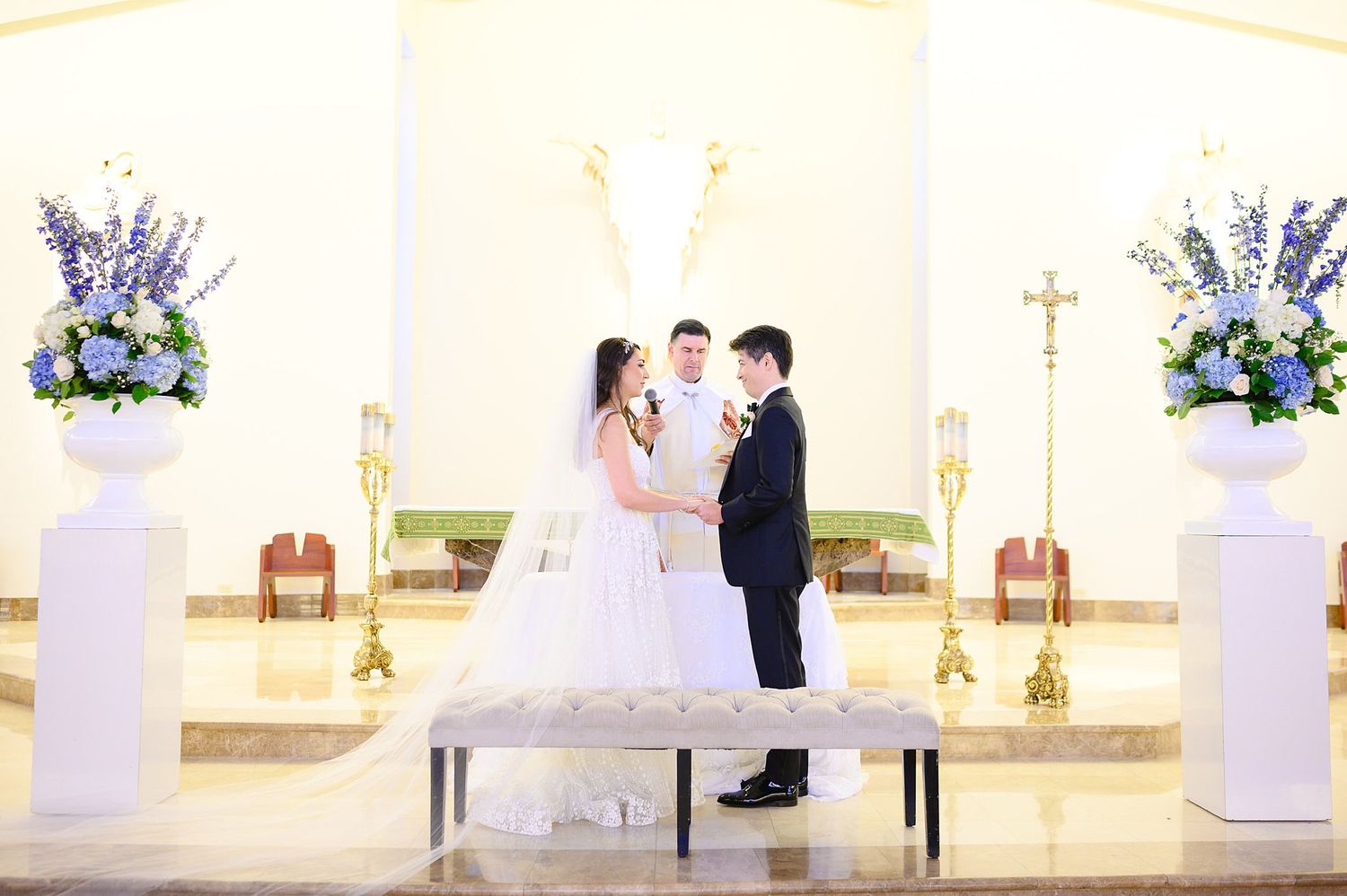 Bride and groom exchange vows at church altar adorned with blue and white floral arrangements.