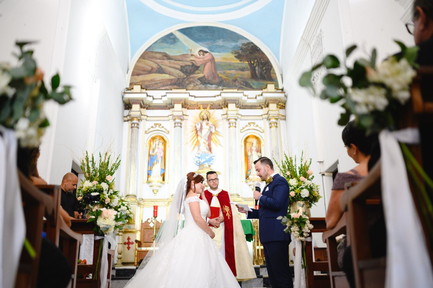Bride and groom exchange vows at a Catholic church altar adorned with white floral arrangements during a wedding ceremony.