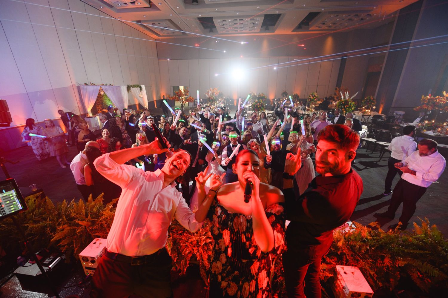 Energetic wedding reception crowd dancing under red lights with guests raising their hands in celebration.