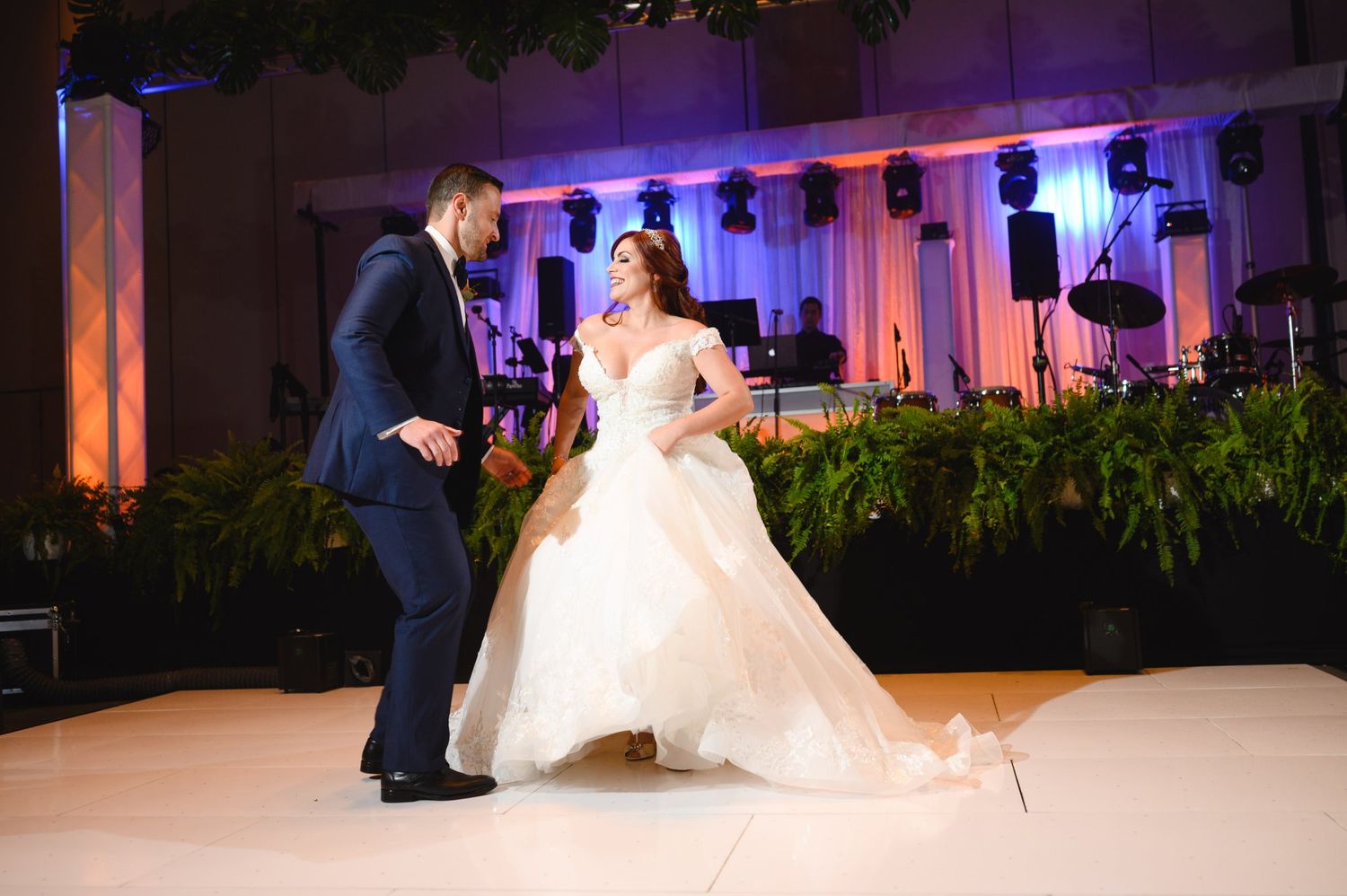 Bride and groom share their first dance at an elegant wedding reception with lush greenery and blue stage lighting.
