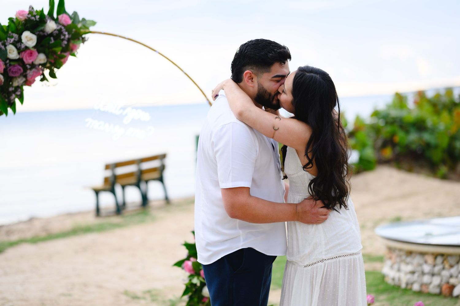 Couple sharing a romantic kiss under a floral hoop arch on a beach during their engagement or wedding ceremony.