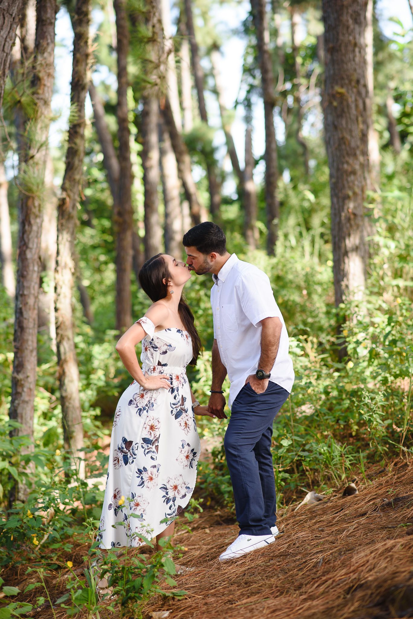 Couple sharing a romantic kiss in a lush forest setting, woman in floral dress, man in white shirt and jeans.
