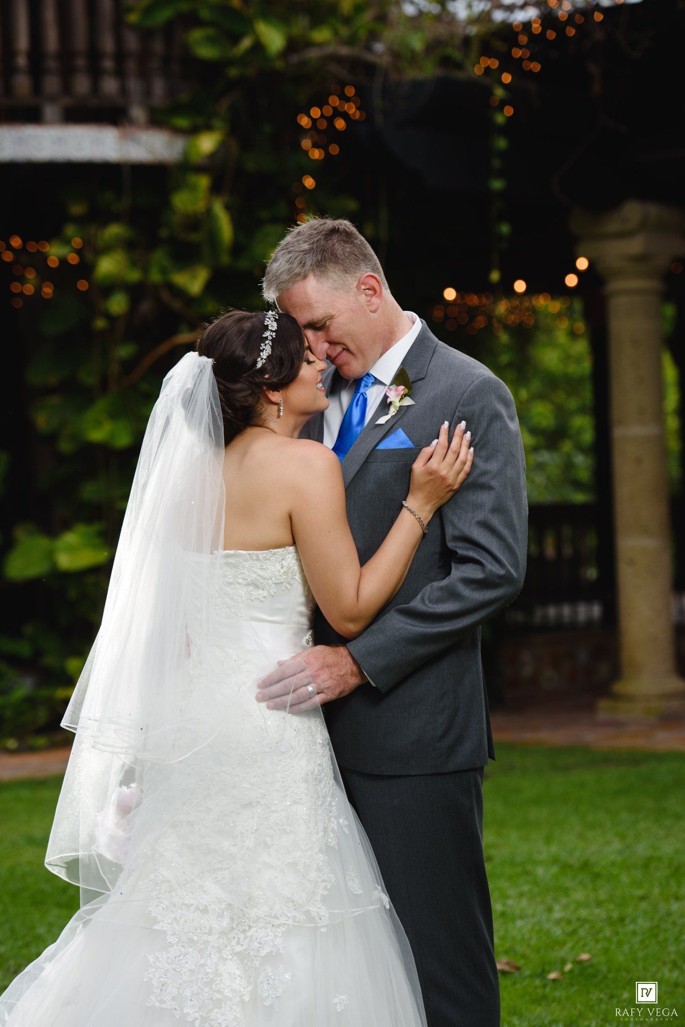 Bride and groom share a romantic embrace on green lawn, she in white lace gown with veil, he in gray suit with blue tie.