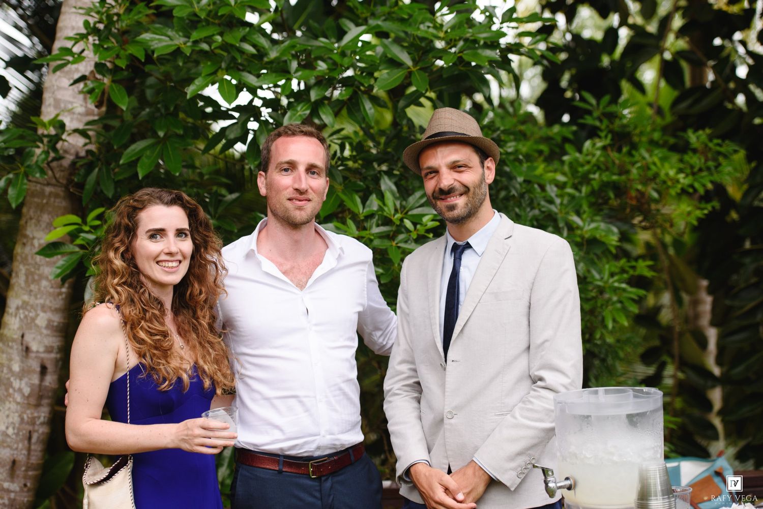 Three guests smile together outdoors at a wedding reception surrounded by lush greenery.