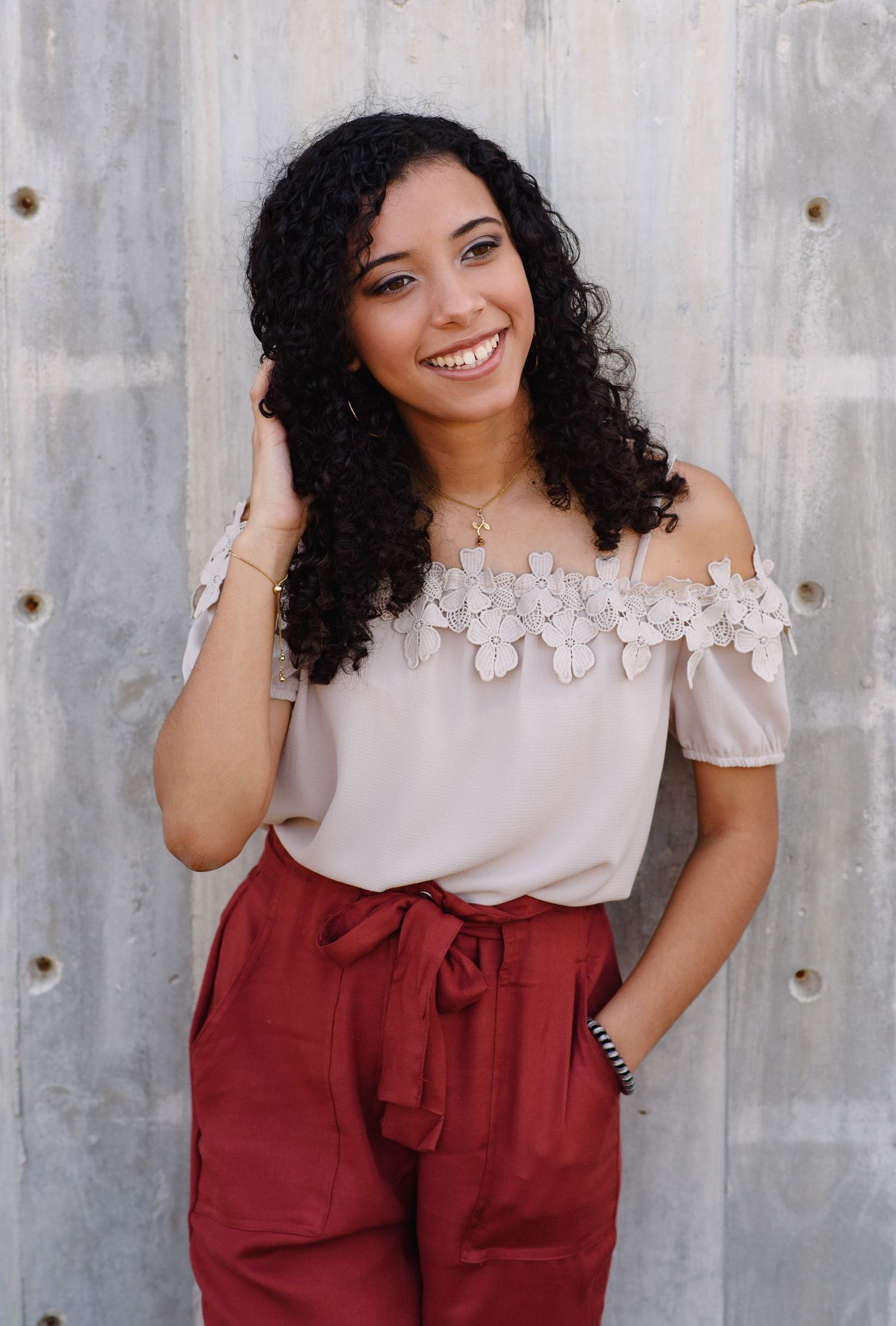 Smiling young woman with curly hair wearing off-shoulder floral lace top and red pants poses against concrete wall.
