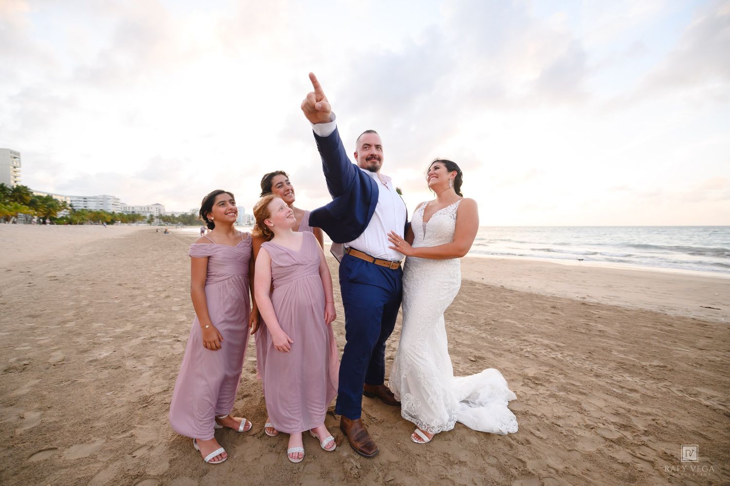 Wedding party posing on a beach at sunset, bride in white gown, groom in navy suit pointing up, bridesmaids in pink dresses.