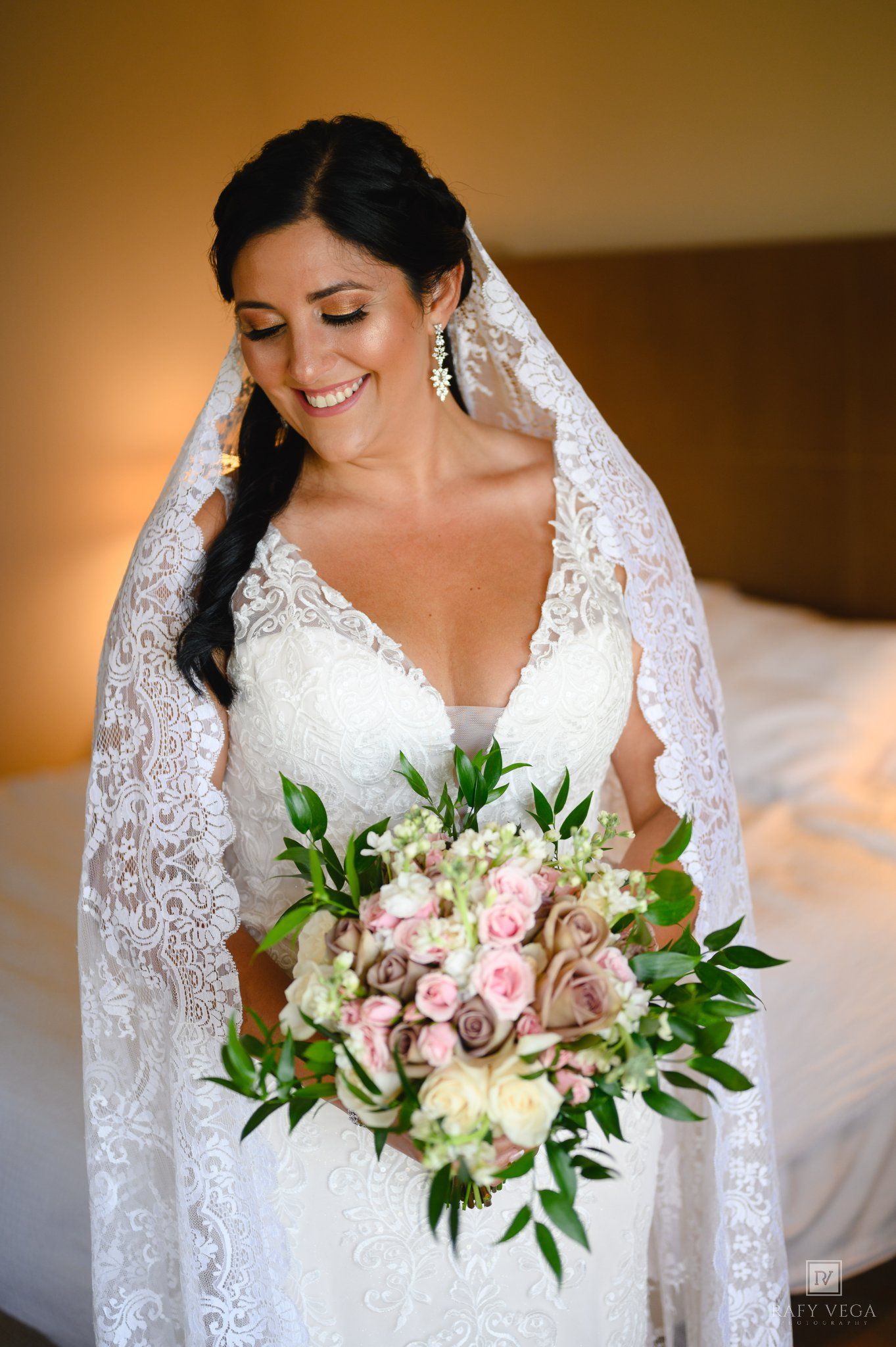 Bride in lace wedding dress and veil smiles holding pink and white rose bouquet with greenery.