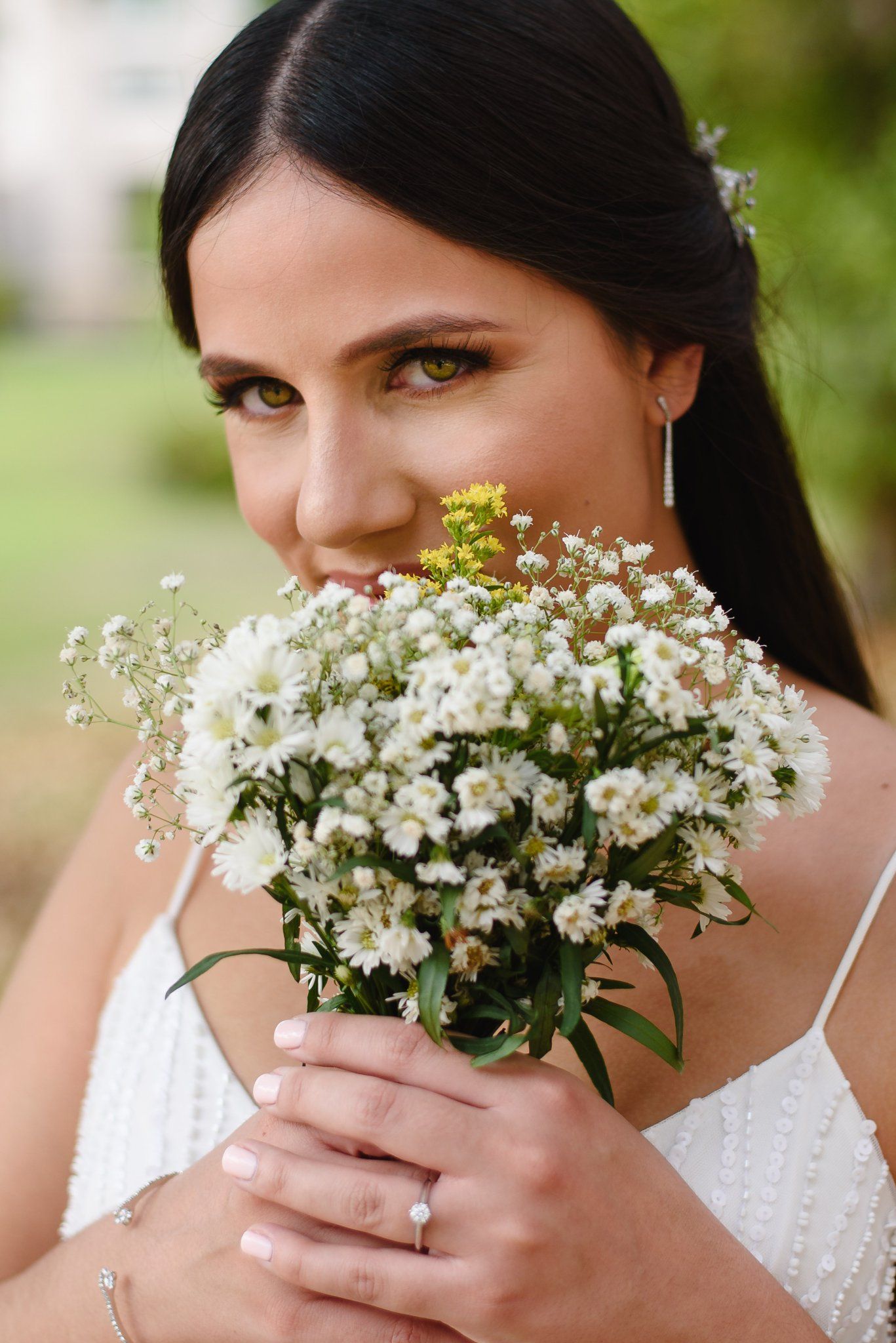 Dark-haired bride holds white wildflower bouquet close to her face, wearing elegant jewelry and white dress outdoors.