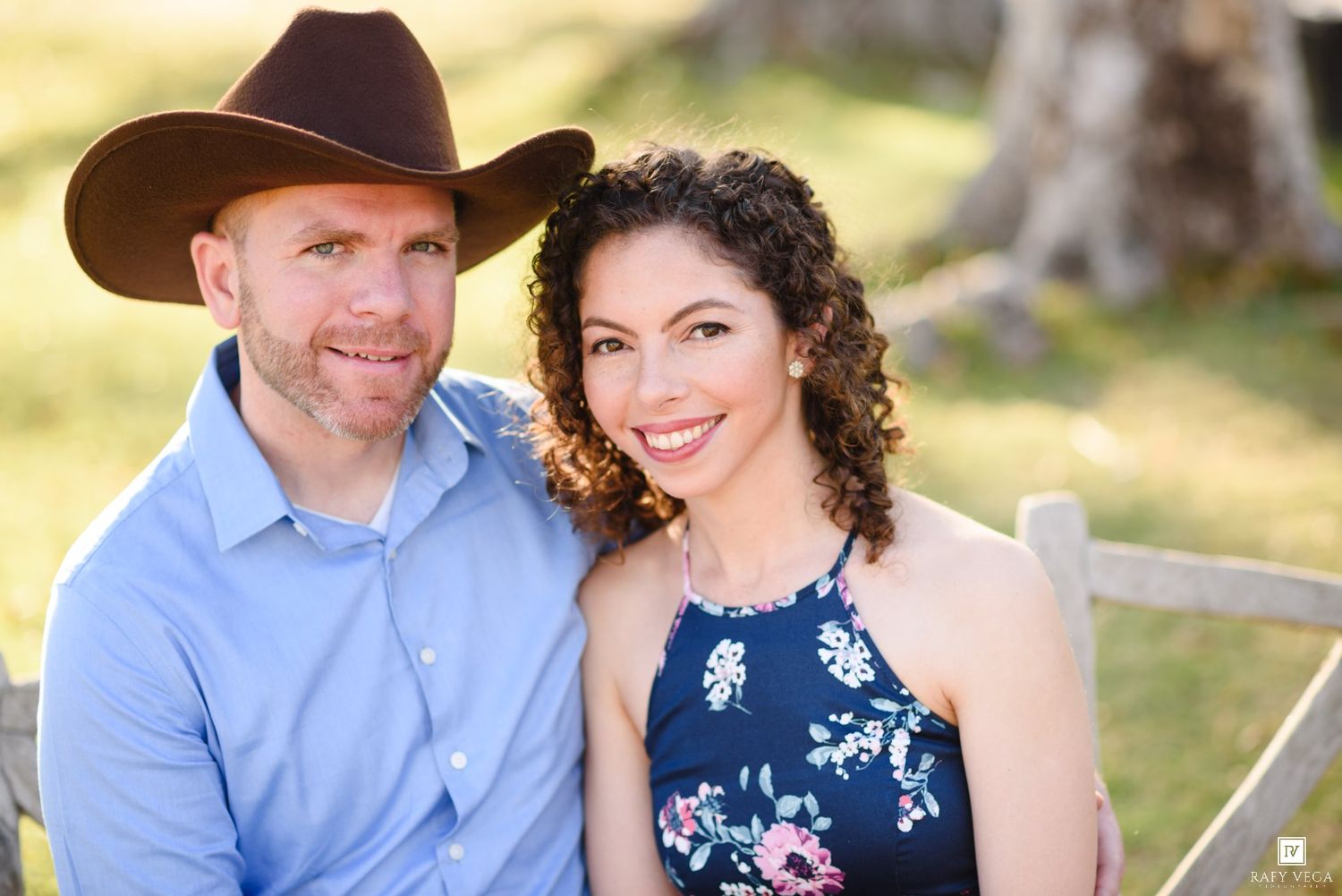 A smiling couple poses outdoors, man wearing a brown cowboy hat and blue shirt, woman in a floral navy dress.