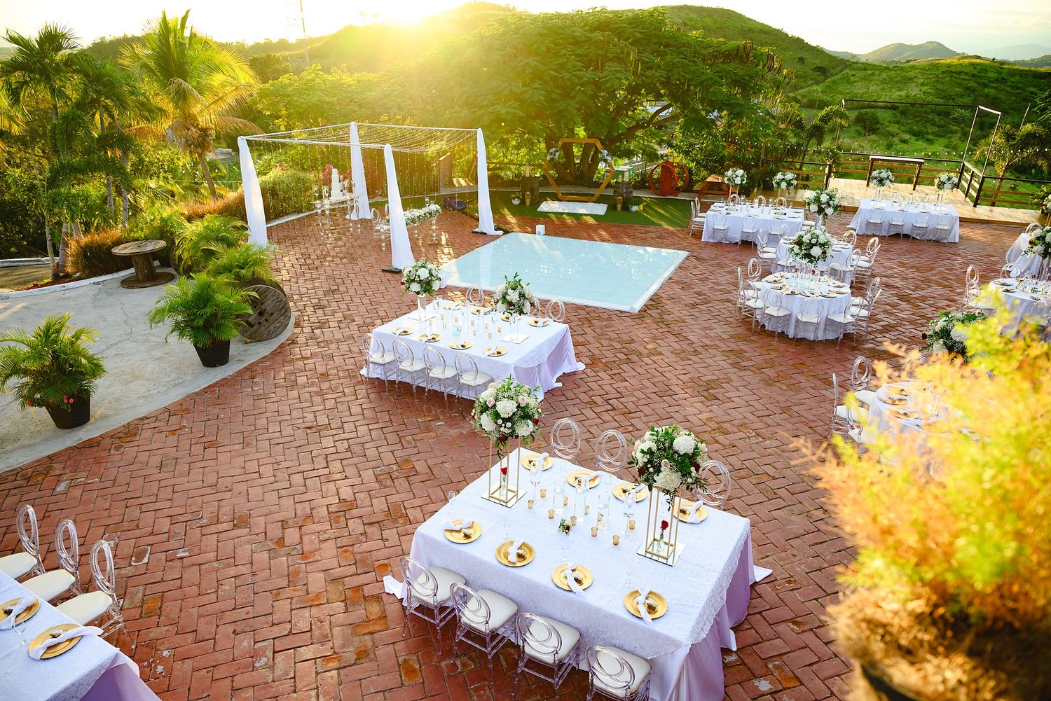 Elegant outdoor wedding reception setup on brick patio with white-draped tables, floral centerpieces, and pool at sunset.