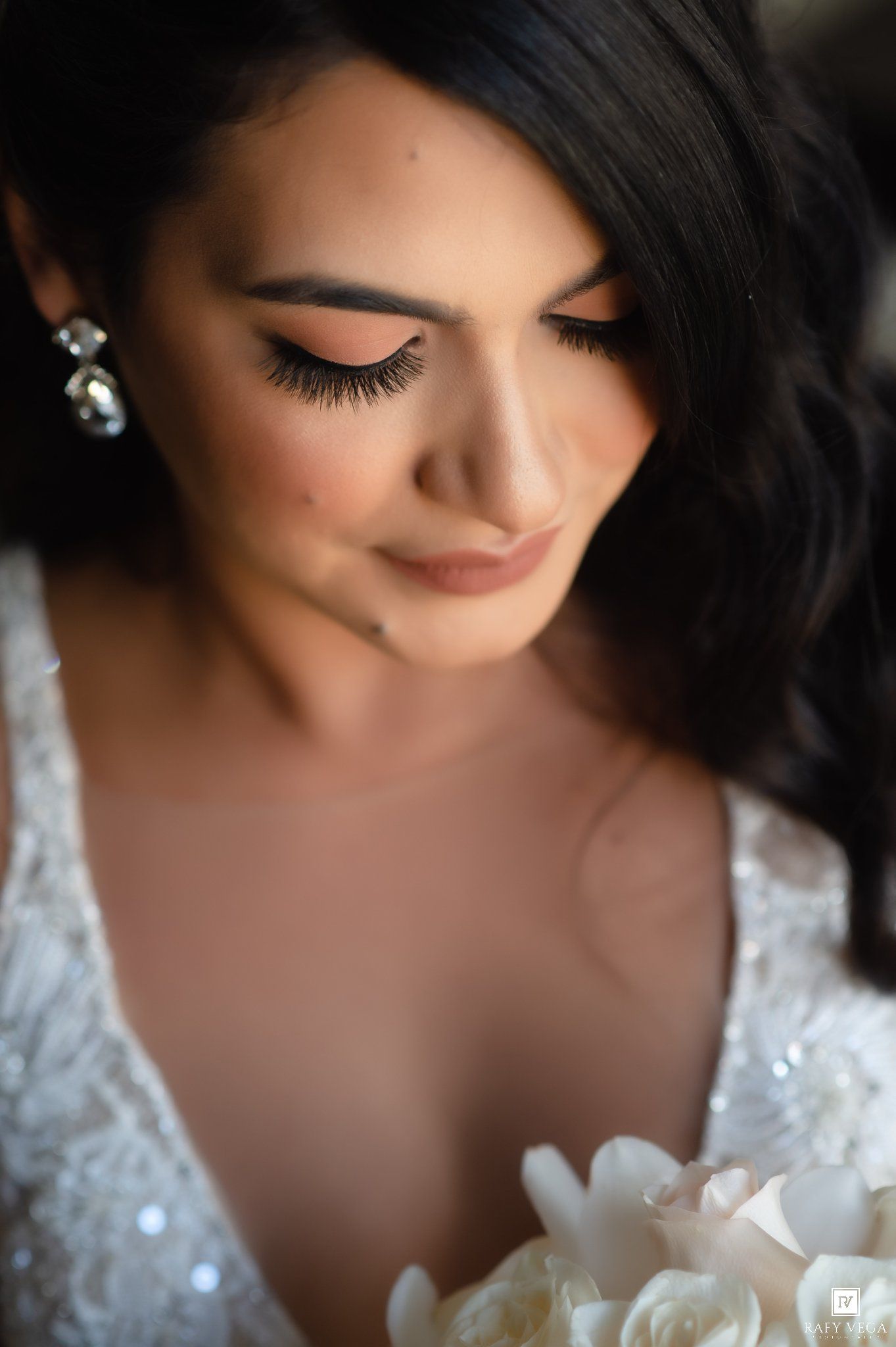 Close-up of a bride with elegant makeup, long lashes, and crystal earrings, looking down at her white floral bouquet.